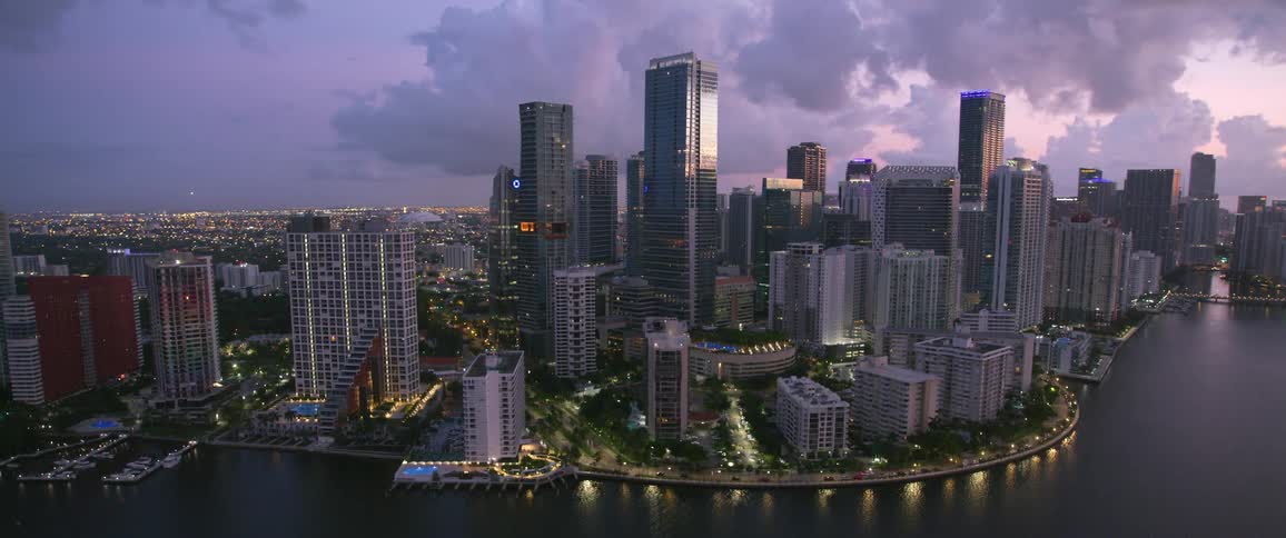 A waterfront metropolitan area with numerous high rise buildings is lit up as the sun has set and it is nearly nighttime