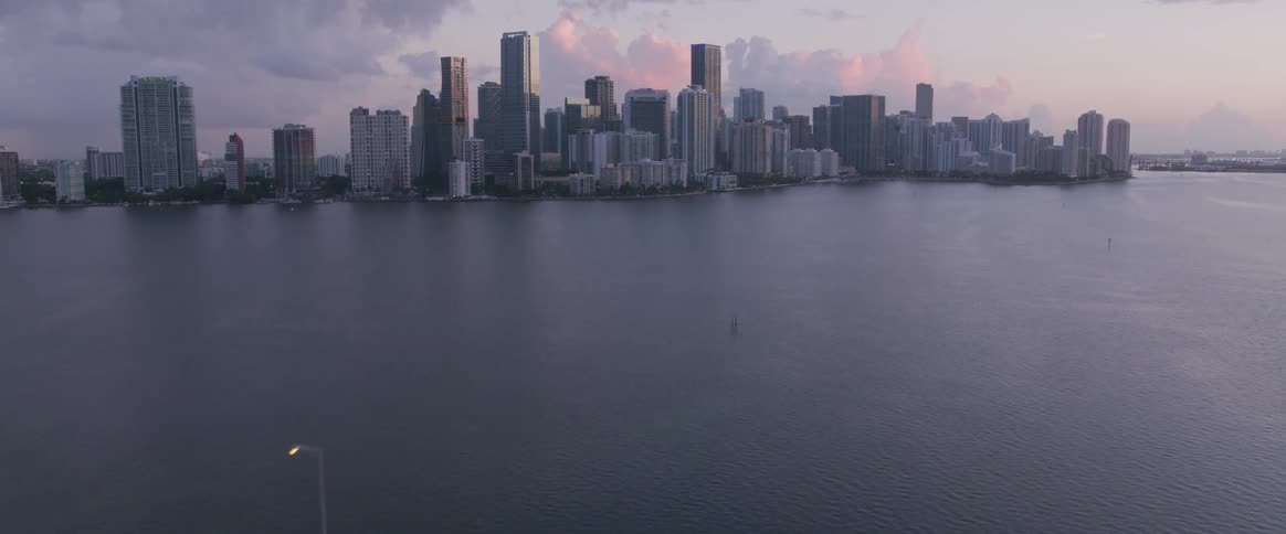 A long bridge can be seen with cars passing over it and the bridge sits on the other side of a large body of water from a metropolitan area