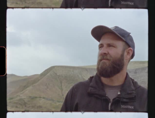 Old film shows a man in a baseball cap standing near a grass covered mountain