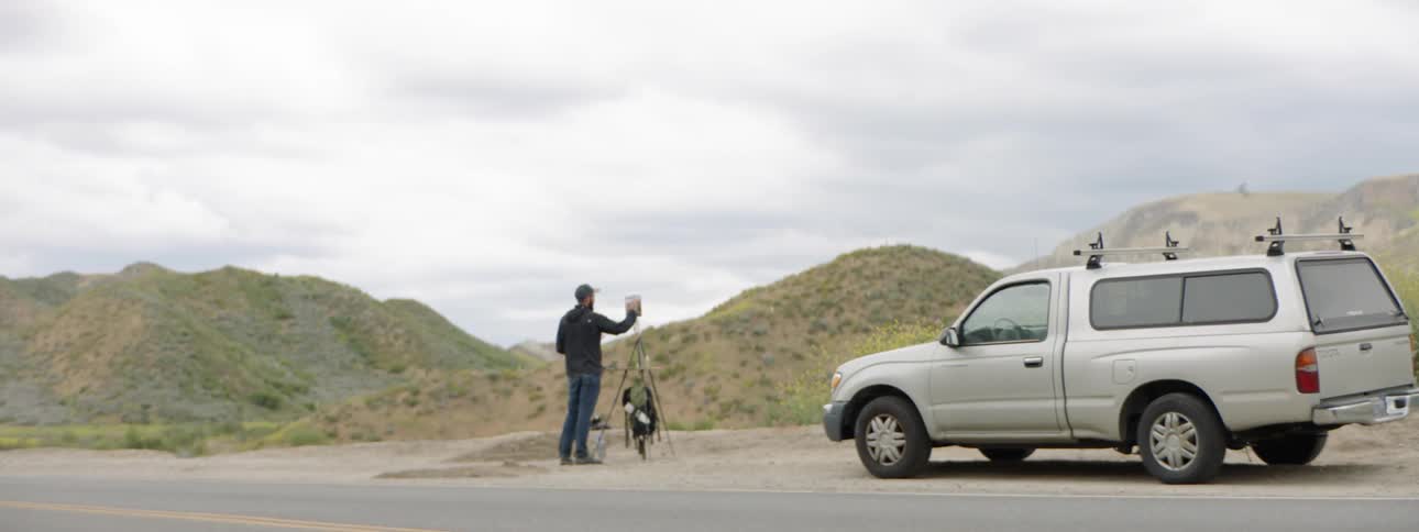 A man walks back toward his easel and canvas he has set up in front of his truck on the side of the road