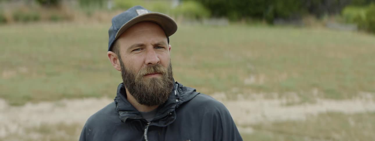 Bearded man wearing cap and coat breathes and looks up on the grass land during daylight
