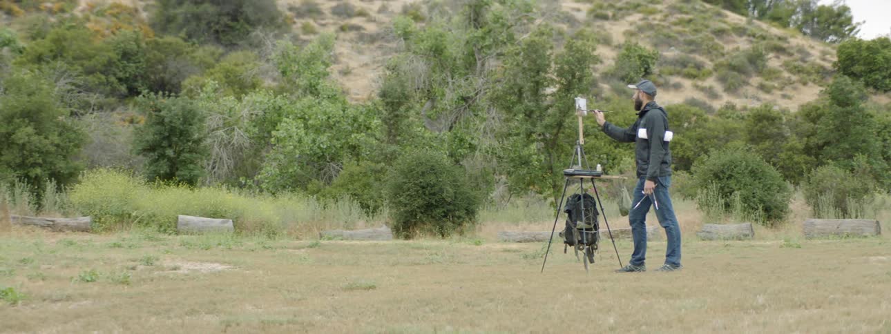 Painter standing in middle of field with easel painting a hillside on canvas
