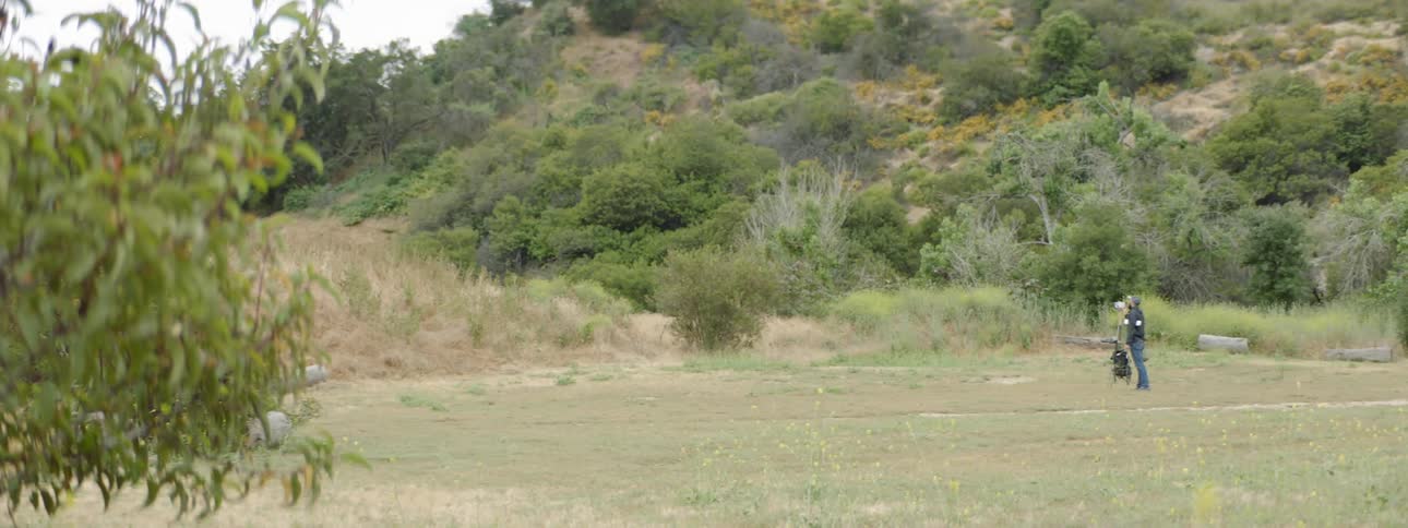A man stands still and looks uphill as he stands in a clearing in a hilly and shrub covered outdoor area