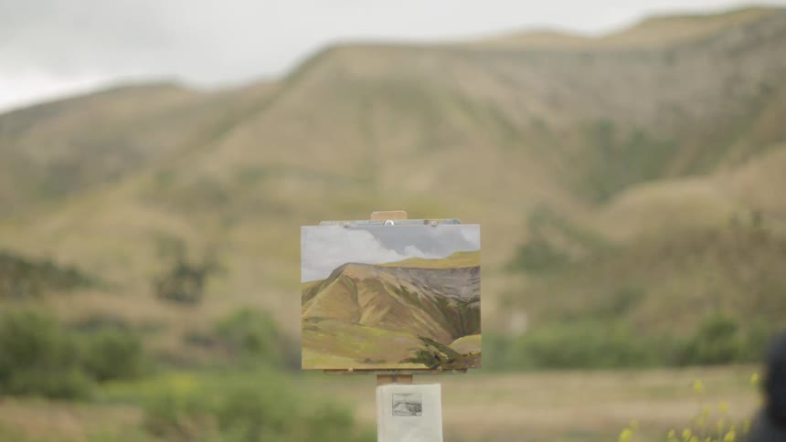 A man stands outside and paints a picture of a hill
