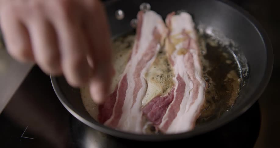 A person's hands place bacon in a hot pan on a stove in slow motion.
