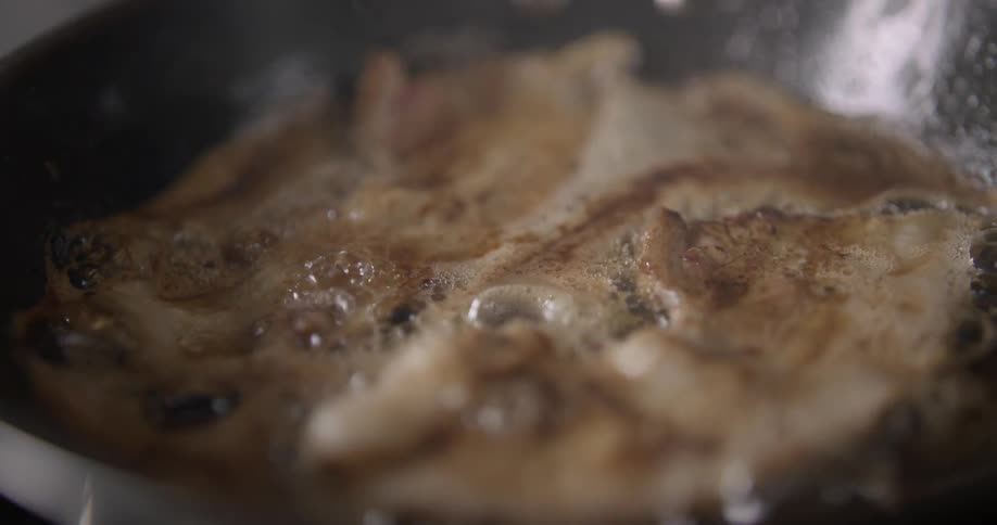 Bubbles and steam emanate from a shaky close-up shot of meat frying in a pan.