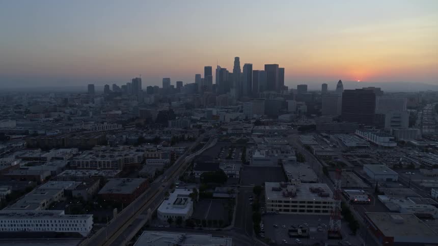 As the sun sits very low in the sky at the edge of the horizon a crowded metropolitan area full of skyscrapers is seen surrounded by a sprawling city
