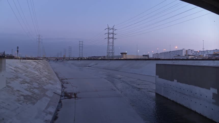 The sky is dusky and the sun is below the horizon as two people can be seen bicycling through the concrete channel portion of the los angeles river and they will soon pass under a bridge