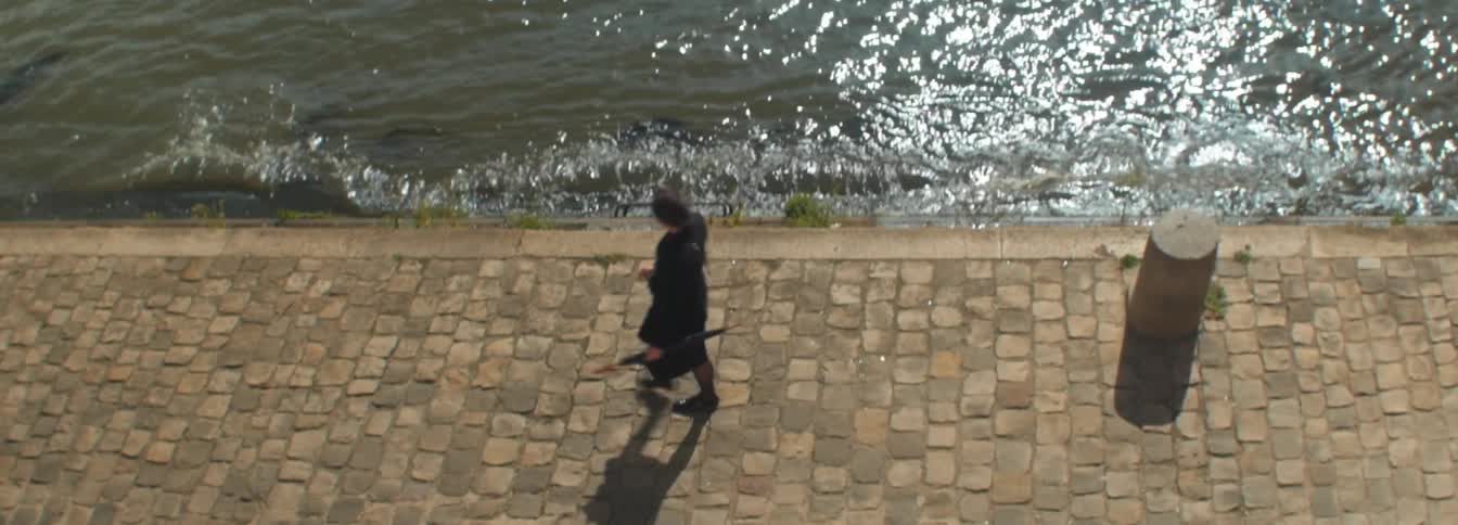 A woman in a black coat carries a closed umbrella while walking on a cobblestone street next to a body of water