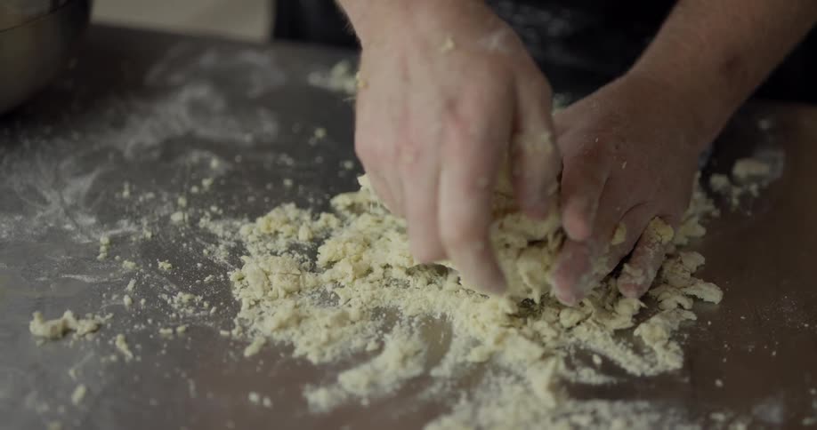 A white man in a black apron kneads dough on a counter in a kitchen