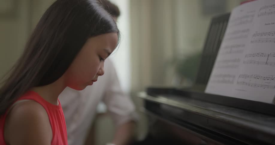 Girl wearing red dress play the piano with the help of man indoors during daylight
