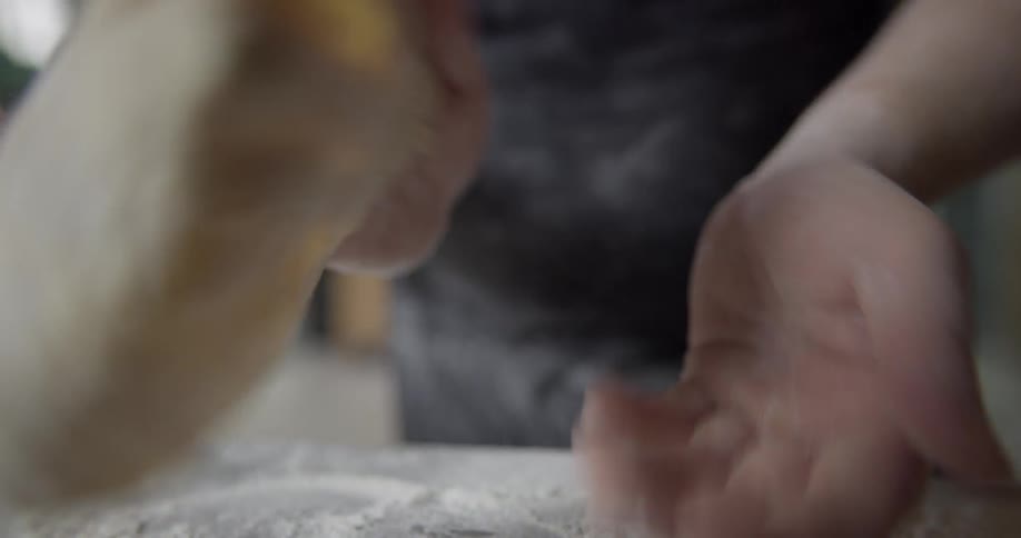 A person's hands can been seen picking up a piece of uncooked dough from a surface covered in flour and then tossing the dough back and forth between their hands to flatten it