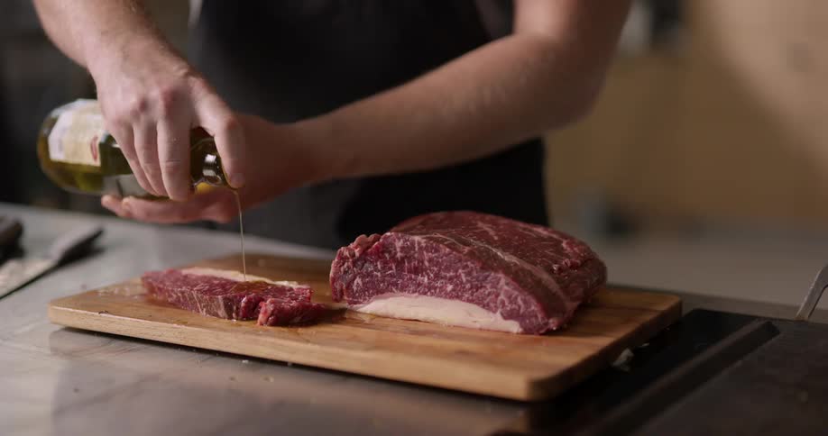 A man pours olive oil on a slice of raw meat and then sprinkles seasoning on top on wood cutting board