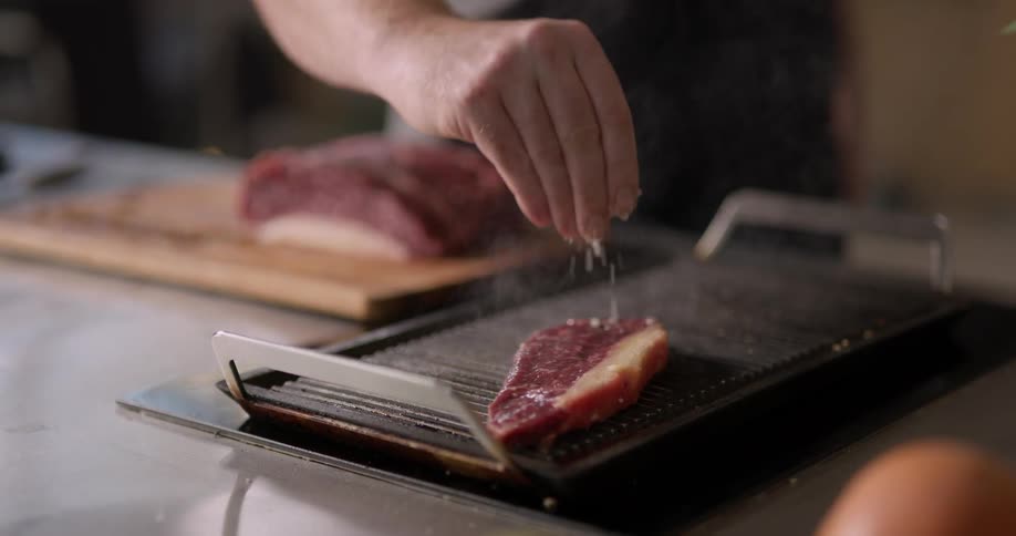 A man takes a slice of beef that he recently cut on a wooden cutting board and places it on a small grill before sprinkling it with salt and letting it cook