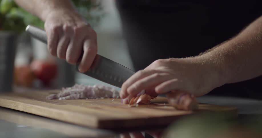 A man chops garlic into very fine slices on a wood cutting board in a kitchen