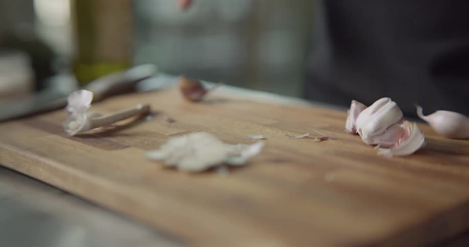 A man slices the ends of a garlic bulb on a wood cutting board and then puts the garlic gloves in a glass jar which he shakes once the lid is on
