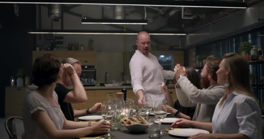 Friends sit and talk at dining table as cook serves dishes