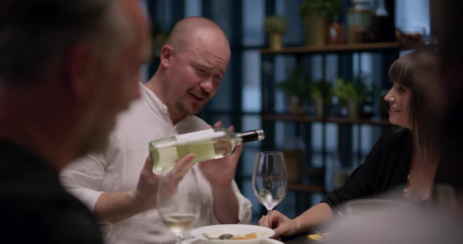 A group of friend sit at the table and man pours wine into glass indoors at dusk