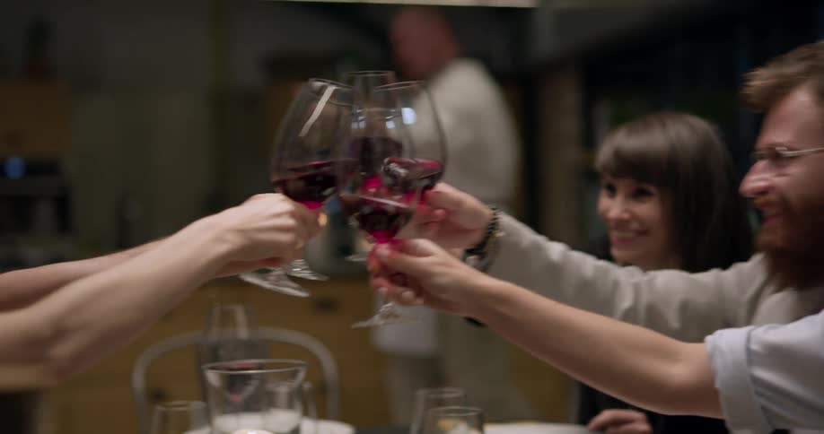 Group of friends sitting at dining table raise their glasses in a toast and laugh indoors
