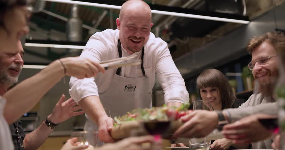 A chef puts down a tray of salad on a table with guests