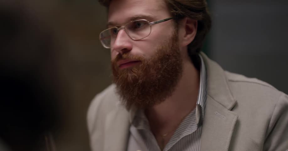 Bearded man wearing glasses sits at the table and eats food at night