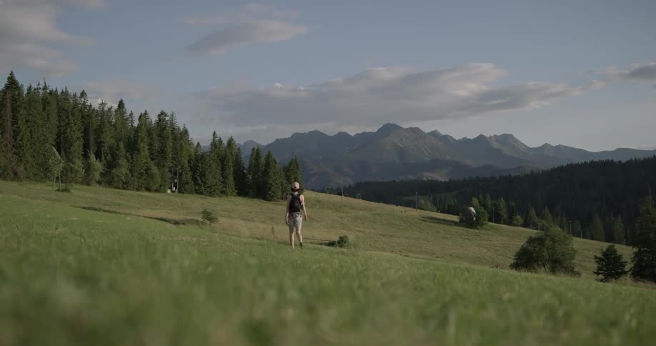 A woman hikes through a field near a forest and mountains