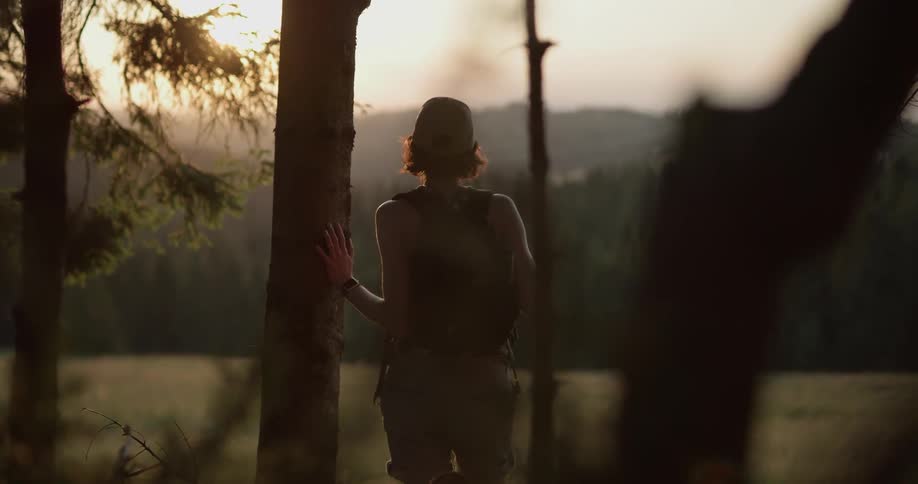 A woman walks out of a forest at sunset