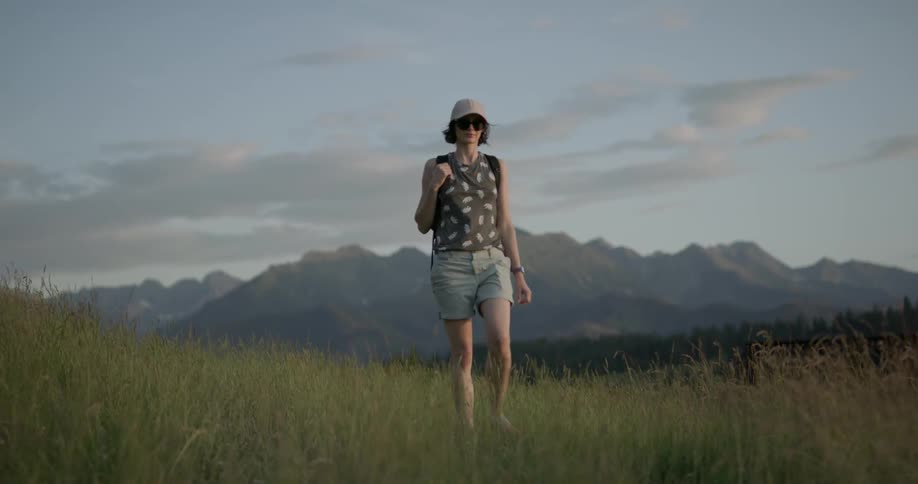 Woman wearing cap and backpack walks on the grassland with the view of distant mountains during daylight