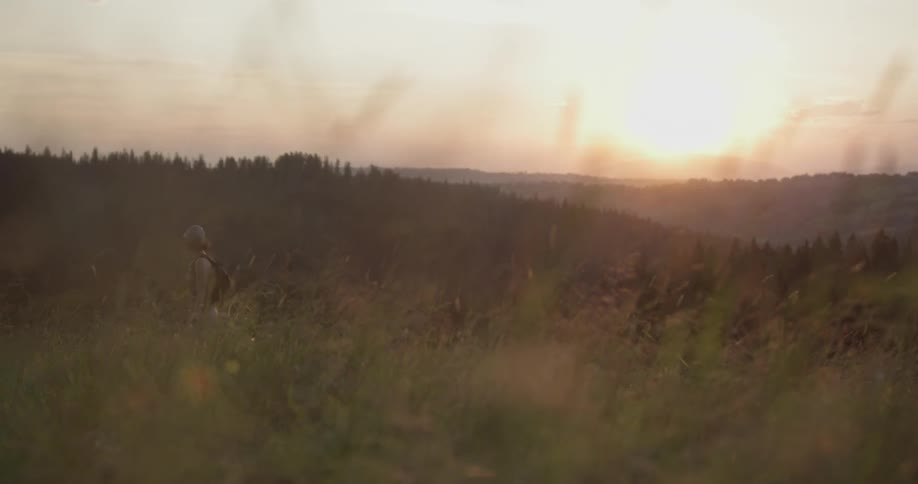 A woman in a hat walks out of a forest into a field at sunset