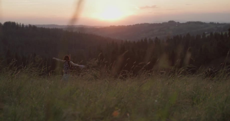 Woman skips through field toward forest and mountains