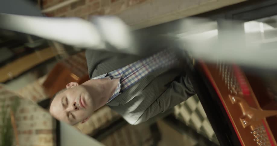 Bald headed man wearing blazer and striped shirt plays the piano indoors