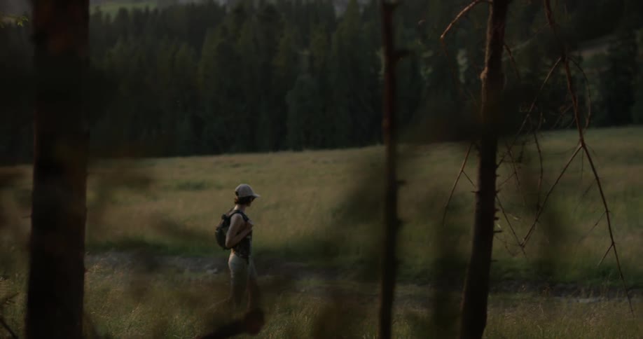 A person wearing a hat and a backpack walks through a field surrounded by forest