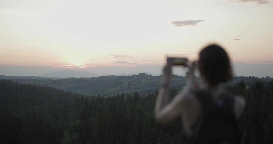 Woman on hiking apparel photographing sunset at the top of a hill