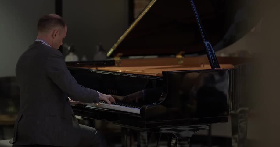 A white man in a gray suit plays a black baby grand piano