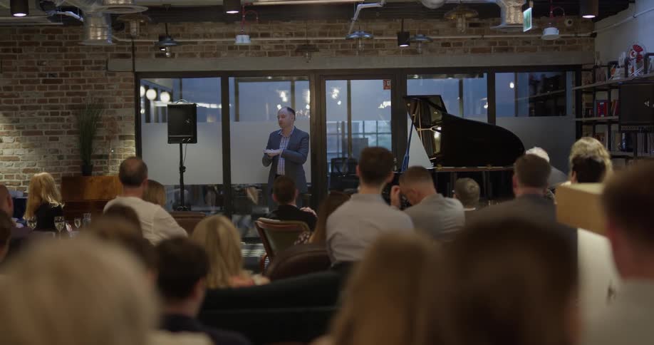 A man stands on a small stage with a piano and speaks to guests