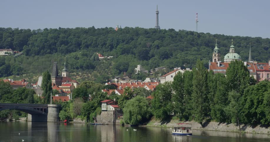 A small boat motors along a calm river at the bank of a town containing several buildings with classical architecture and domes