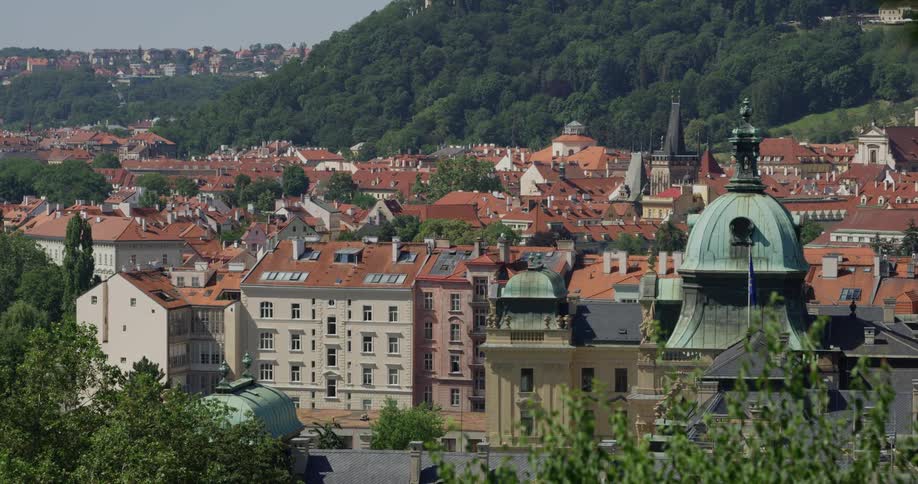 A european city sits at the base of a large forest covered hill and the town is full of many multistory buildings made with distinctive architecture