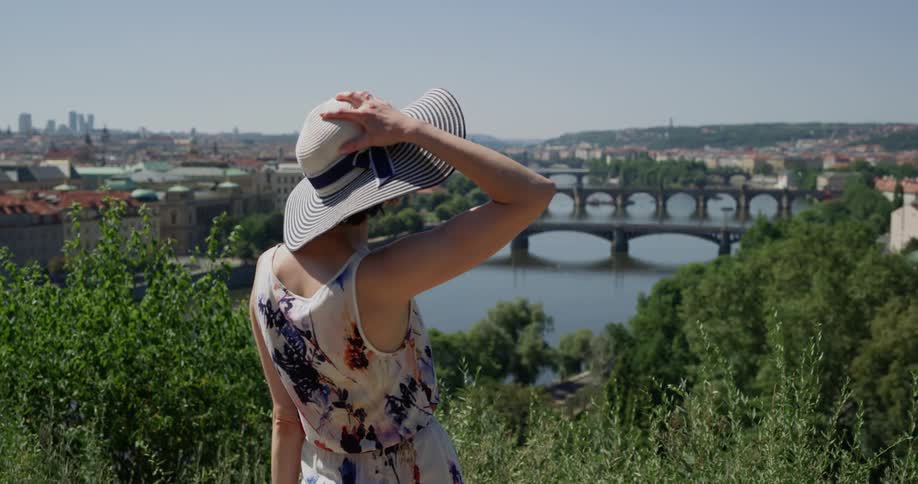 A woman in a sun hat walks to a clearing atop a hill that overlooks a river that is spanned by several bridges with very nice architecture that connect parts of a european town