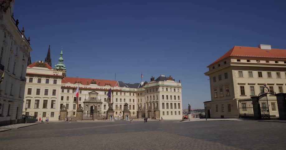 A plaza in the center of several buildings with red roofs and nice architecture has a handful of pedestrians walking through on a clear and sunny day
