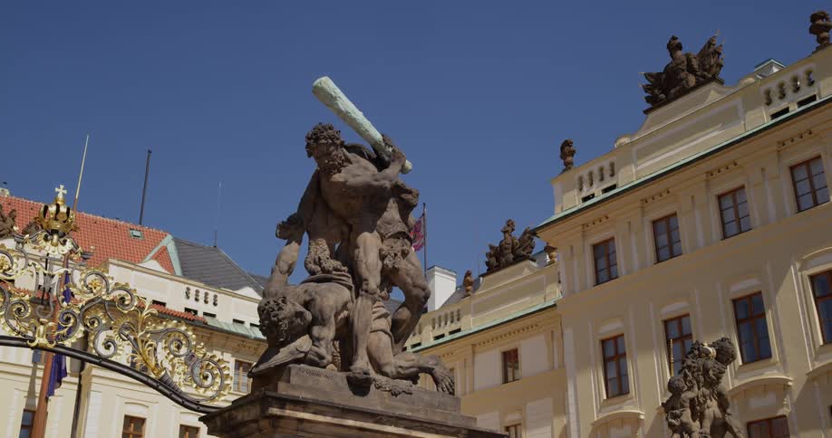 In the middle of several ornate buildings with classical architecture there is a sculpture of a muscular man holding a club over another man who has fallen down