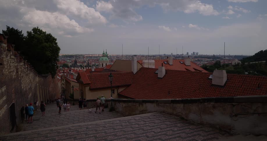 Several people are walking down a paved pathway with multiple sets of stairs in it which leads alongside some buildings in a densely packed European city with a mix of modern and older architecture
