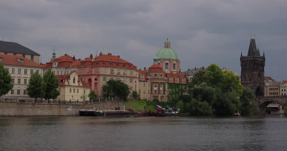 Boats are docked along the bank of the vltava river near the charles bridge with the dome of the church of st francis visible