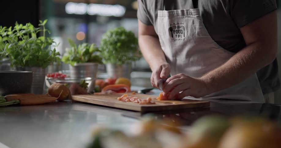 A man wearing an apron is carefully slicing food with a knife on a cutting board that is located on a counter top surrounded with several potted herb plants