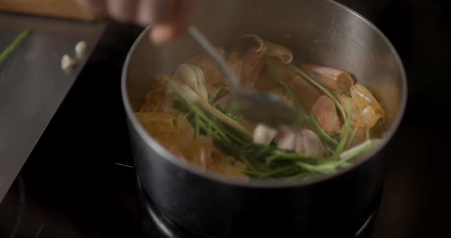 A steel pot is simmering atop a stove and it is filled with a soup base vegetables and shrimp that a person is stirring with a metal spoon