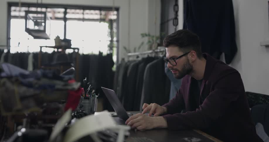 A man sits at a desk working on a laptop in a store with clothing on racks