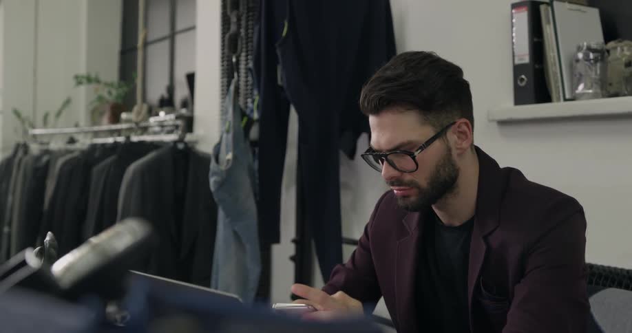 Man wearing glasses and blazer sitting at the table uses smartphone in the clothing store during the day