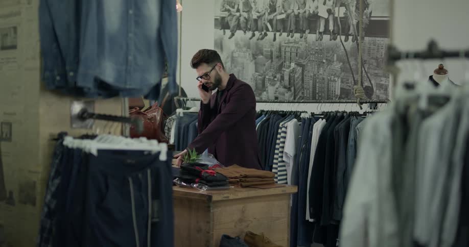 A man in a clothing store talks on a cell phone while looking at pants then walks away from the pants while still talking on the phone