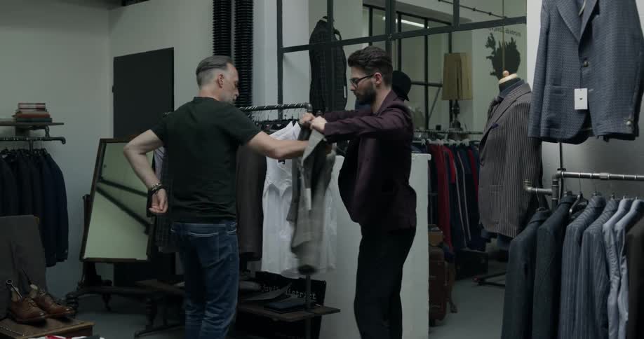A salesperson helps a man with gray hair put on a gray suit jacket and then hangs the hanger on the clothing rack