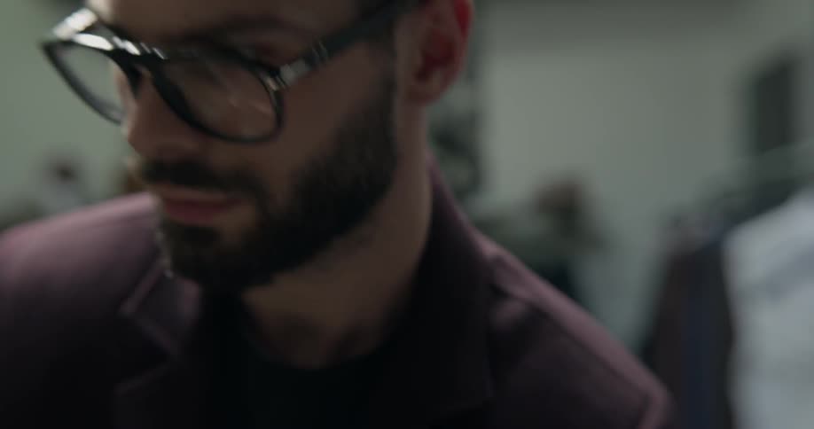 A bearded man wearing glasses and a suit coat thoroughly inspects a piece of clothing in a menswear store that has several mannequins in suits on display in the background