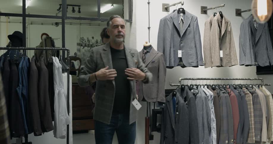 A clothing store professional removes a jacket from a hanger from a clothing rack and helps another man try the jacket on